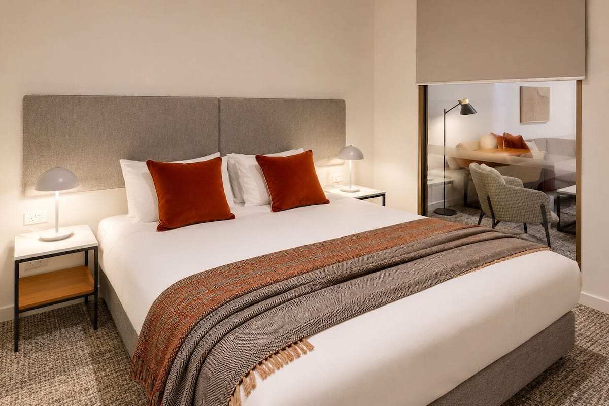 Hotellihuone, jossa on suuri sänky, kaksi yöpöytää, työpöytä, tuoli ja lasioven kautta avautuva näkymä oleskelutilaan.