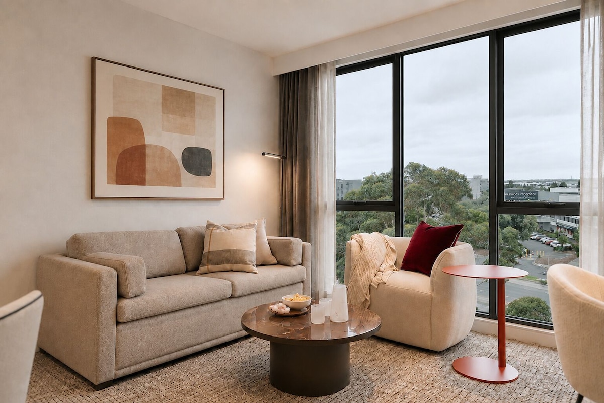 A modern living room with a sofa, armchair, and coffee table, a large window with a view, and a framed abstract painting.
