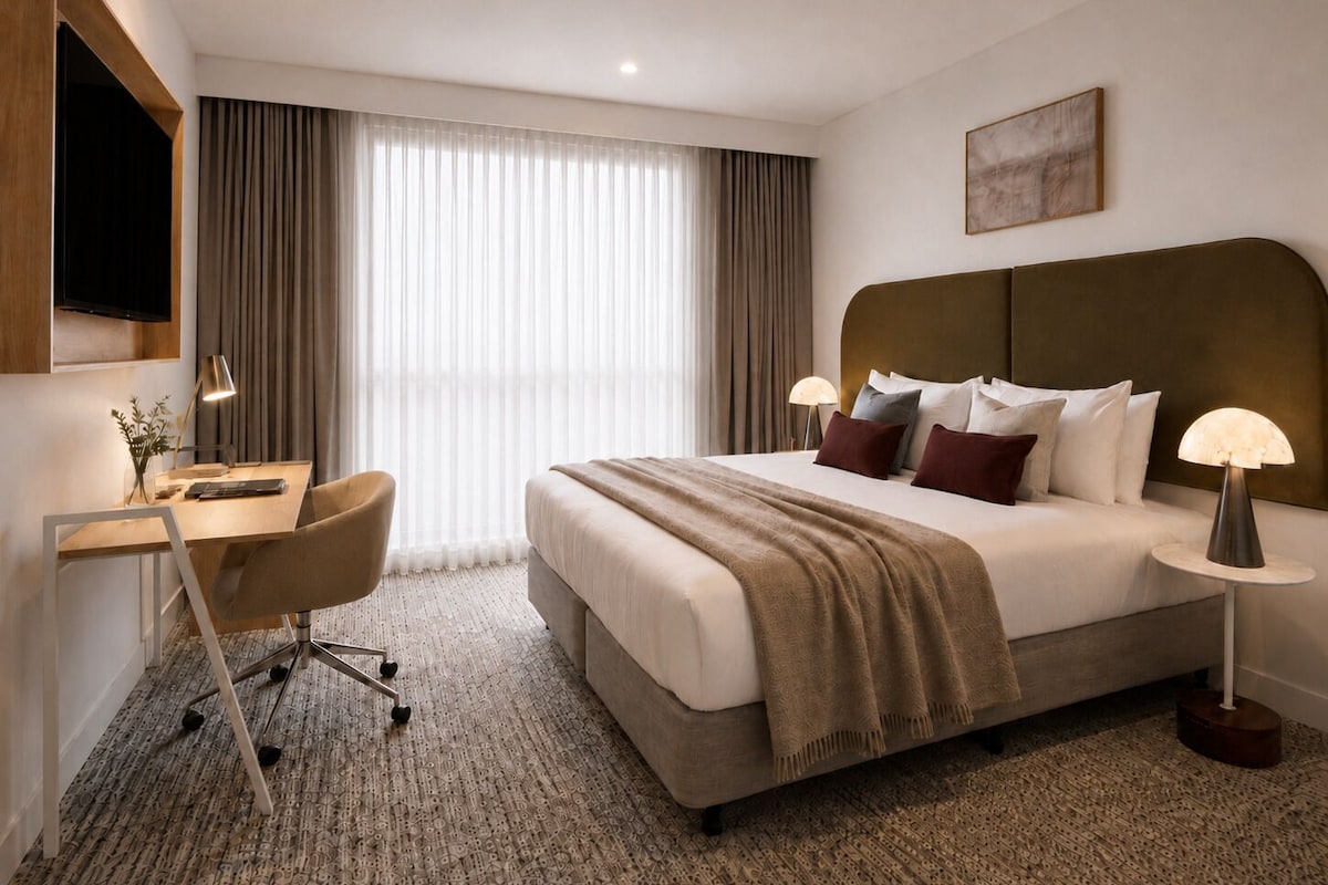 A hotel room with a bed, desk, chair, and a large window with curtains.