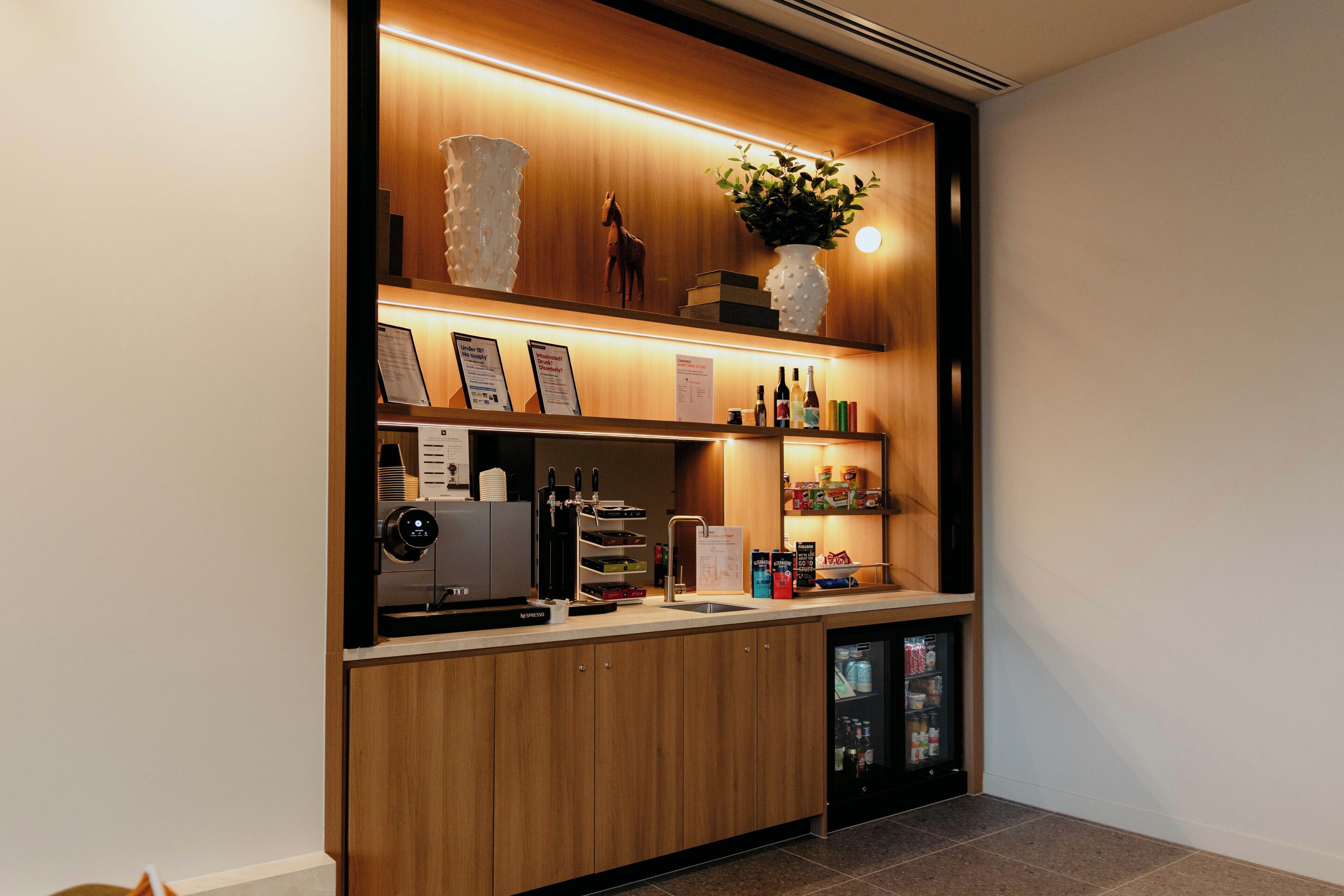 A modern bar area with a coffee machine, shelves with bottles, a refrigerator, and a decorative plant.