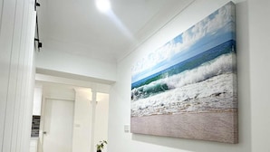 Un pasillo con piso de madera, paredes blancas y un cuadro grande de una escena de playa.