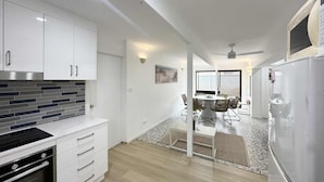 Cocina moderna con armarios blancos, placa de cocina negra y horno empotrado. Hay un comedor con mesa y sillas, y un refrigerador a la derecha.