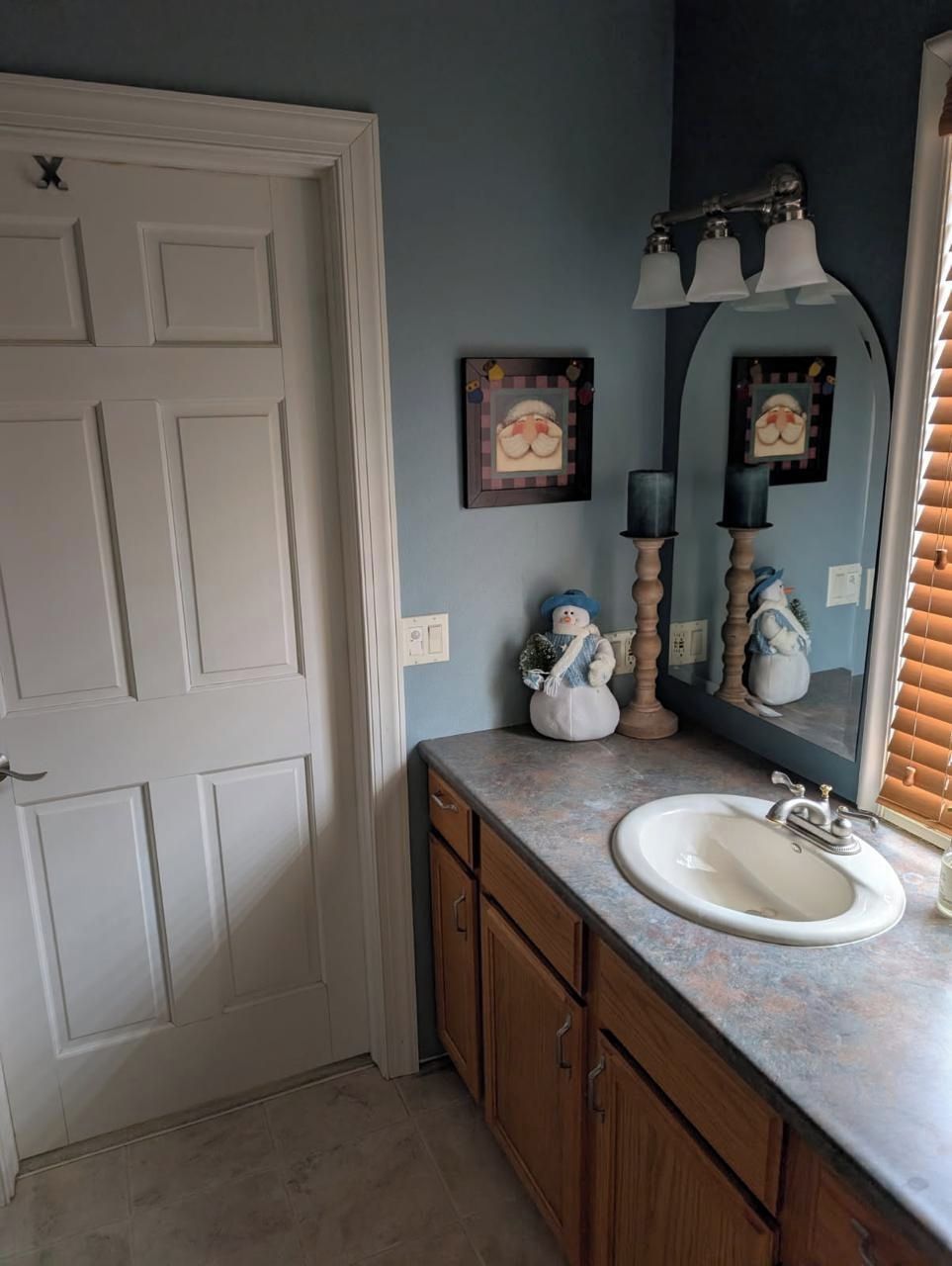 A bathroom with a white door, a sink, wooden cabinets, a mirror with three lights, and two framed pictures on the wall.
