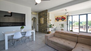 Ein modernes Wohnzimmer mit einem beigefarbenen Sofa, ein Essbereich mit weißem Tisch und Stühlen, eine Küche mit Steinwand und ein großes Fenster mit Blick.
