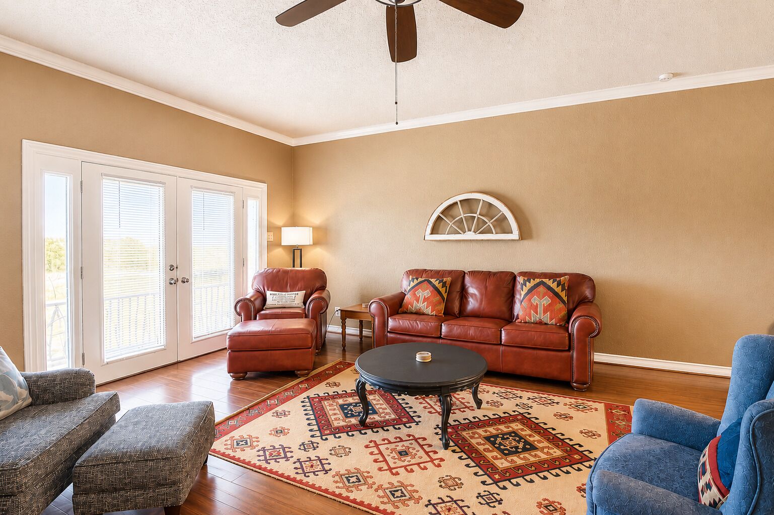 Un salon avec un canapé en cuir marron, un fauteuil bleu, un tapis à motifs et un ventilateur de plafond.