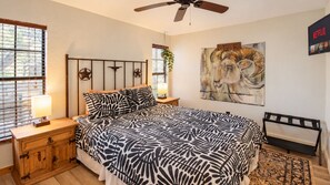 Une chambre à coucher comprenant un lit, des tables de chevet, un ventilateur de plafond, un tableau, un téléviseur et une fenêtre avec des stores.