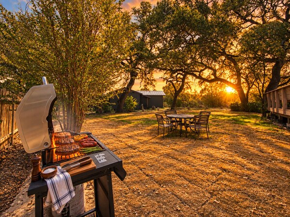 An outdoor barbecue setup with a grill, a table, and chairs, surrounded by trees and a sunset in the background. - Newly renovated rustic home with deck by Horseshoe and Tubing (Canyon Lake)