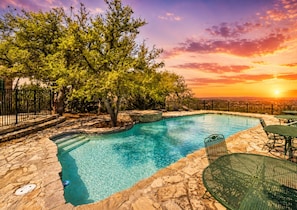 A swimming pool with stone tiles, surrounded by trees and a sunset view.