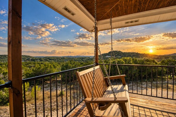 A wooden swing chair on a balcony with a metal railing, overlooking a landscape with trees and hills. - Spacious house with living room, kitchen, balcony and Hill country views (Canyon Lake)