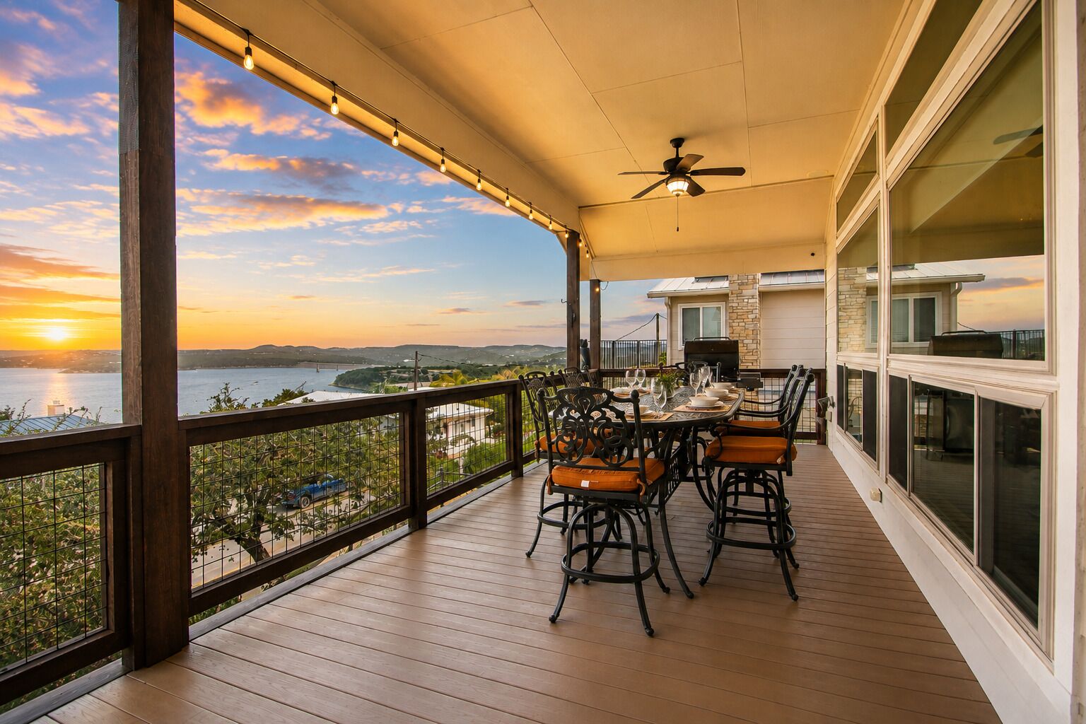 A covered outdoor patio with a table set for dining, overlooking a body of water and a cityscape during sunset.