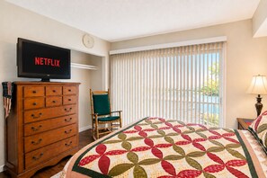 A hotel room with a bed, a wooden dresser, a television displaying 'NETFLIX', a chair, and a window with blinds.