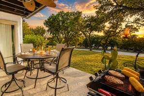 An outdoor dining area with a table set for four, surrounded by chairs, and a grill with food being cooked.