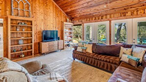 Un interior acogedor de madera con un sofá, televisión, estanterías y un ventanal grande.