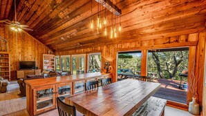 Una cabaña de madera espaciosa con una gran mesa de comedor, múltiples sillas, un televisor y una terraza con vistas a los árboles.