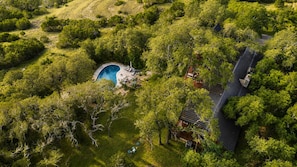 An aerial view of a property with a swimming pool surrounded by lush greenery, a traditional-style building, and a curved roof.