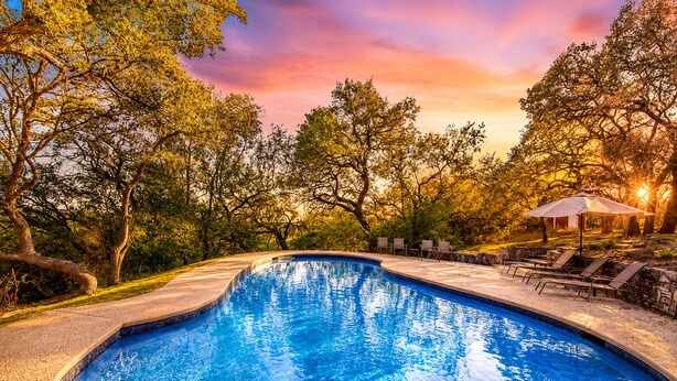Una piscina de borde curvo, rodeada de árboles y un área de descanso con sillas y sombrillas.
