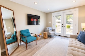 A living room with a teal armchair, a grey armchair, a beige sofa, a flat-screen TV mounted on the wall, and a large mirror. - Lovely home on 2+ acres with views, firepit & outdoor kitchen - wedding perfect (Dripping Springs)