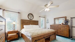 Un dormitorio con cama de madera, mesita de noche, cómoda, ventilador de techo y ventana con cortinas.