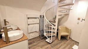 Een compacte badkamer met een witte wastafel, toilet en een witte handdoekhouder. Er staat een beige stoel in de buurt van het toilet en in de achtergrond is een wenteltrap te zien.
