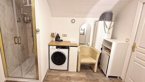 Een kleine badkamer met een douche, wasmachine, stoel en een klein kastje.