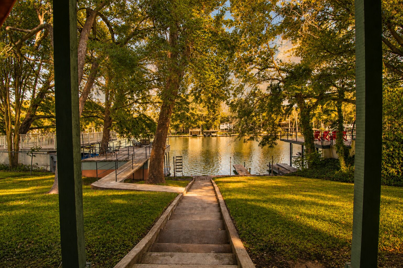 A park with a concrete pathway leading to a dock over a calm lake, surrounded by trees and a fence.