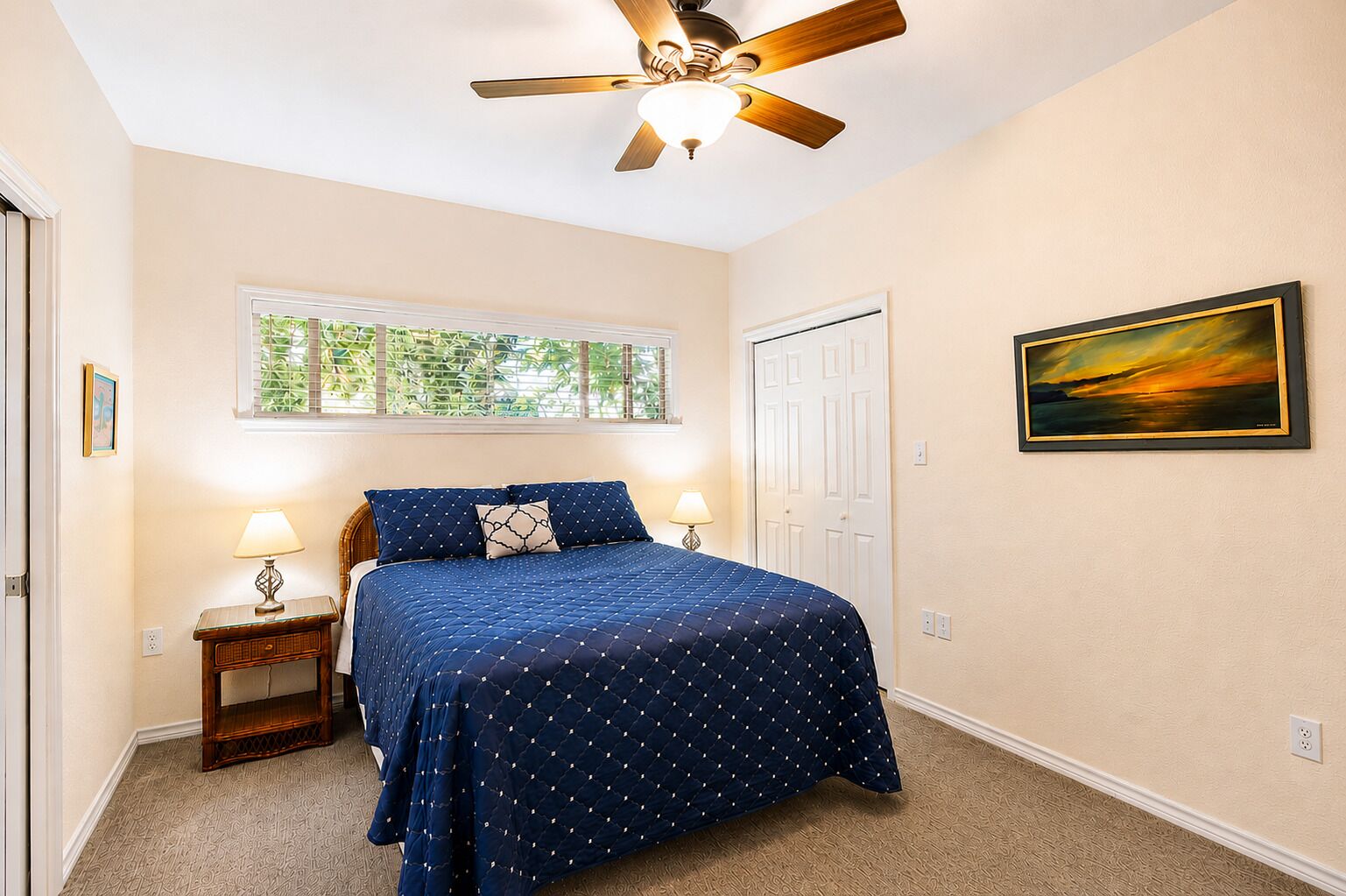 A bedroom with a bed, two bedside lamps, a ceiling fan, a window with a view of greenery, and a framed picture on the wall.