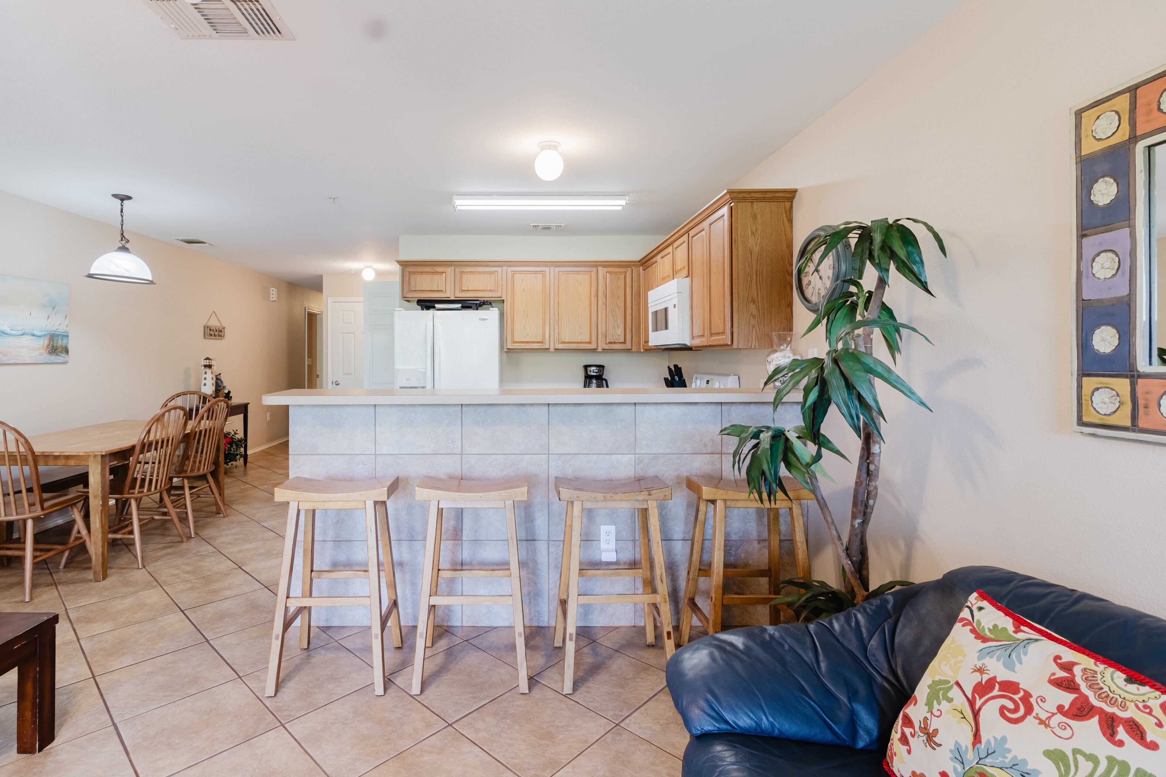 A kitchen with wooden cabinets, a white refrigerator, a microwave, and a coffee maker. There are three wooden stools and a dining table with chairs. A large potted plant is in the corner.