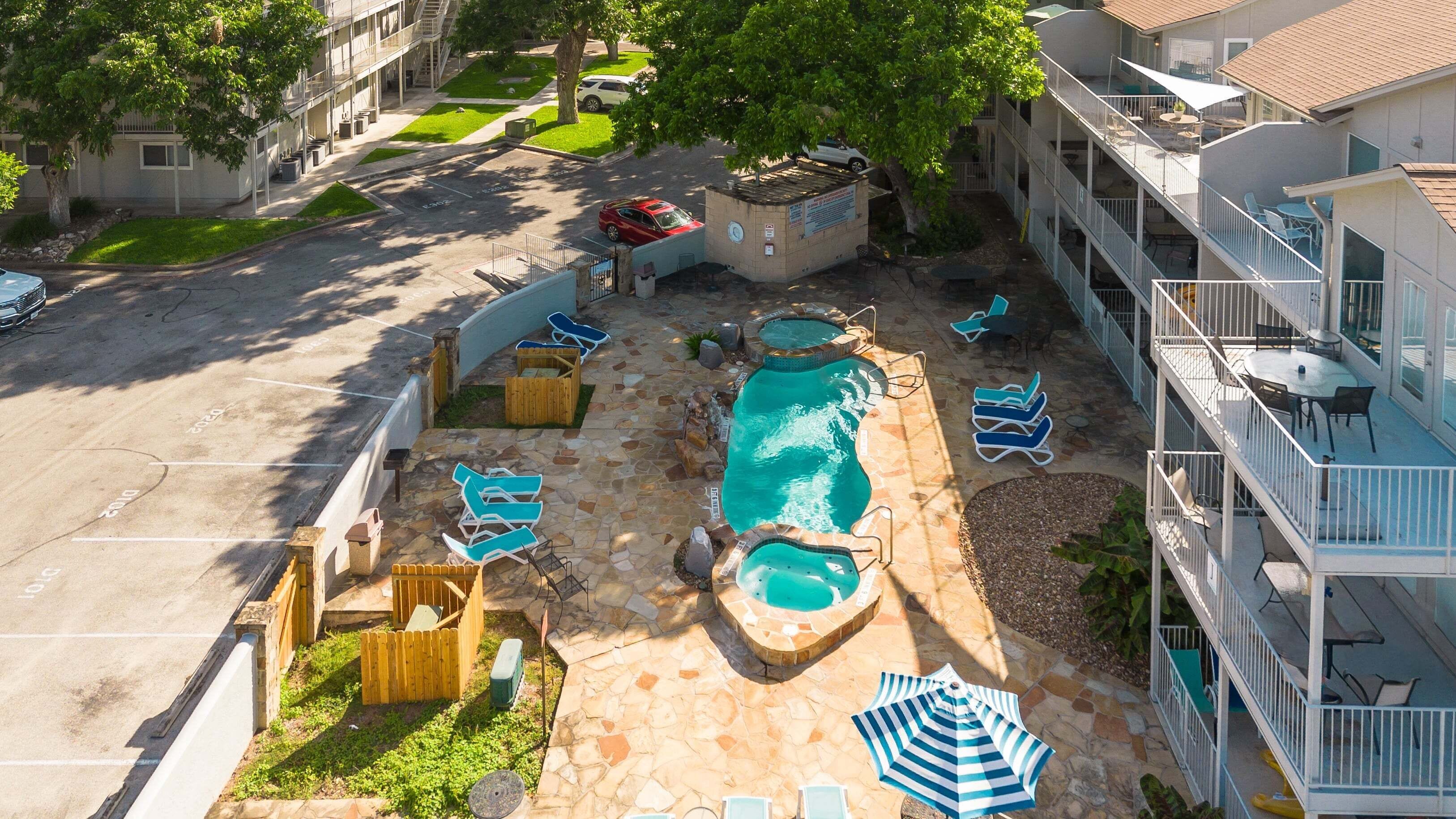 A swimming pool with lounge chairs and umbrellas, a parking lot, and a multi-story building with balconies.