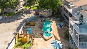 A swimming pool with lounge chairs and umbrellas, a parking lot, and a multi-story building with balconies. - 3BR | 5 min Drive to Schlitterbahn | Pool, Hot Tub (New Braunfels)