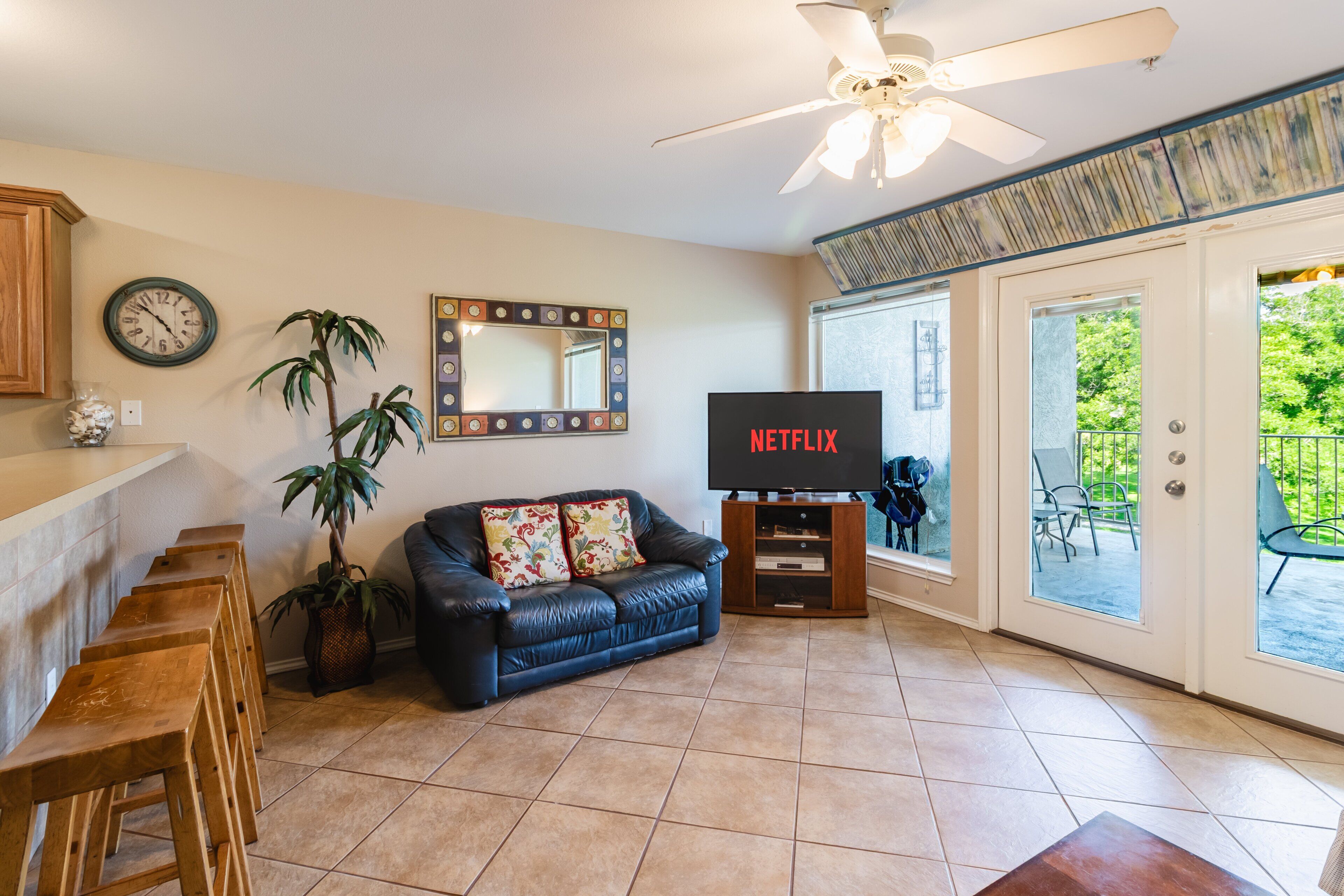 A living room with a black leather sofa, wooden stools, a television, a ceiling fan, a clock, and a large mirror.