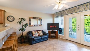 A living room with a black leather sofa, wooden stools, a television, a ceiling fan, a clock, and a large mirror. - 3BR | 5 min Drive to Schlitterbahn | Pool, Hot Tub (New Braunfels)
