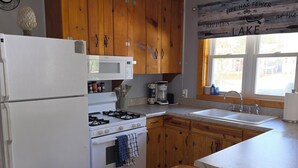 Een keuken met houten kastjes, een witte koelkast, een fornuis, een magnetron en een raam met uitzicht op de omgeving.
