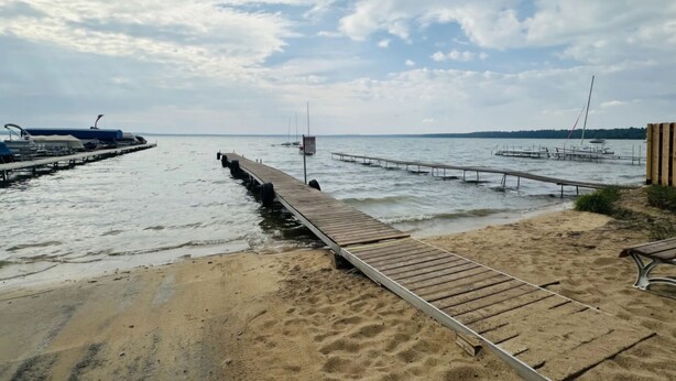 Een strand met een houten pier die zich uitstrekt over kalm water, een zandstrand en een verre kustlijn met boten.