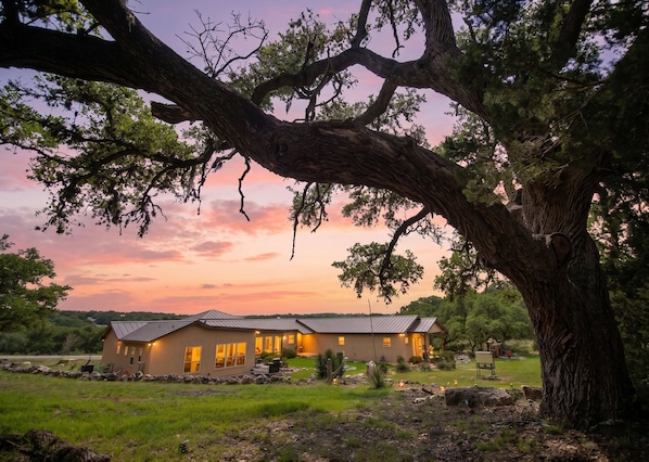 A house with a large tree in the foreground, a well-maintained lawn, and a clear view of the surrounding landscape. - Steps to Remi's Ridge & Hayes Hollow Century Oak Casita - Near Canyon Lake (Spring Branch)