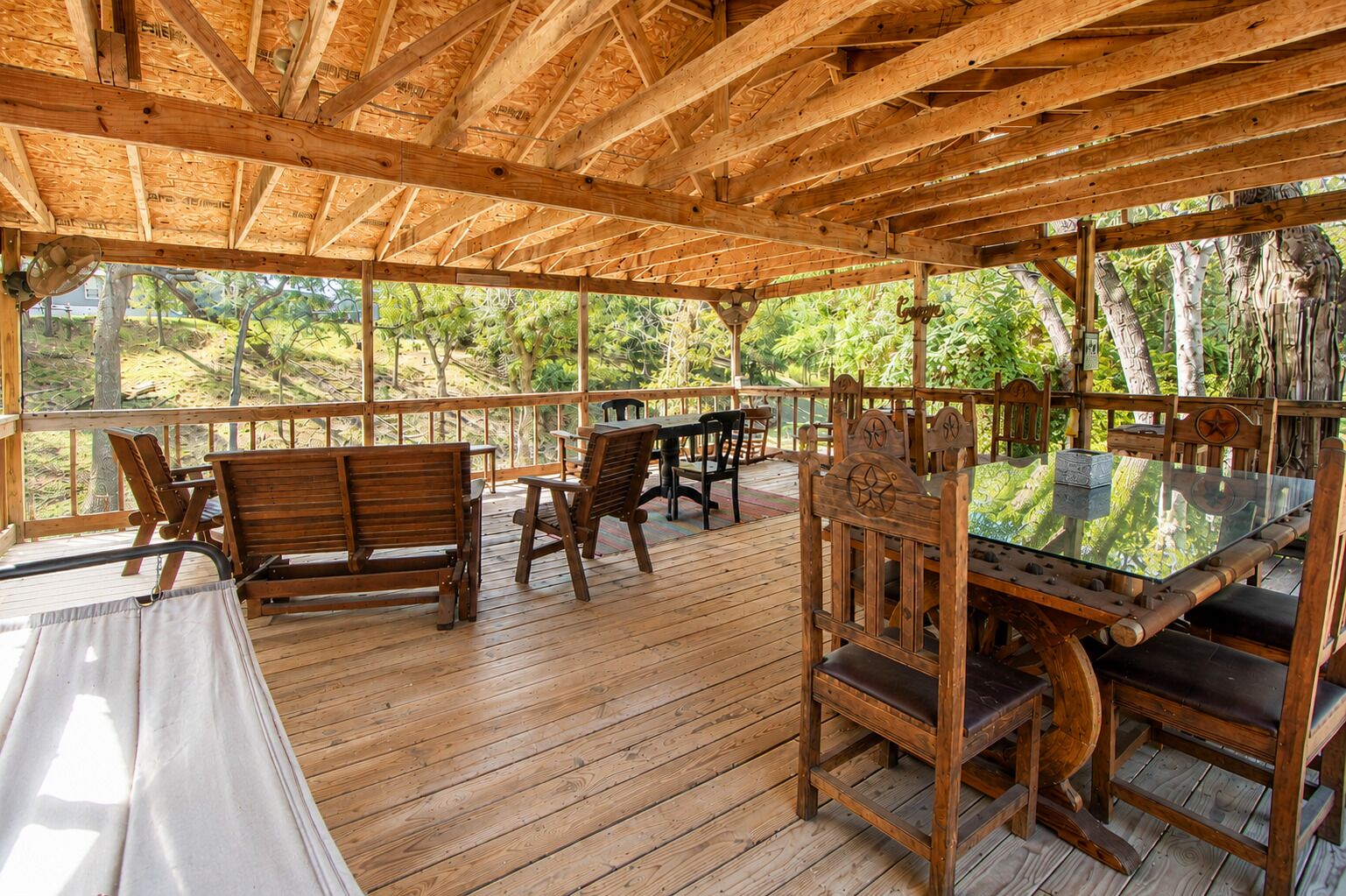 An outdoor wooden deck with a glass table and chairs, surrounded by trees.