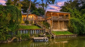 A wooden cabin with a balcony overlooking a body of water, surrounded by lush greenery. - Charming dog-friendly home with expansive deck - near boat ramp & attractions (New Braunfels)