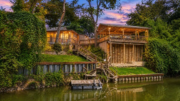 Une cabane en bois dotée d’un balcon donnant sur une étendue d’eau, le tout entouré d’une végétation luxuriante.
