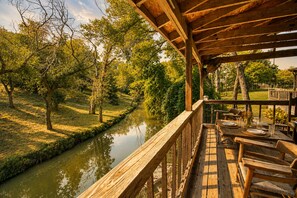 Une terrasse en bois avec une table dressée pour le repas, offrant une vue sur une rivière paisible et une végétation luxuriante.