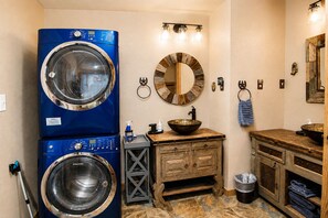 Un lavadero con lavadora y secadora de carga frontal color azul, un mueble de tocador de madera con un espejo grande y un lavabo con un cuenco decorativo.