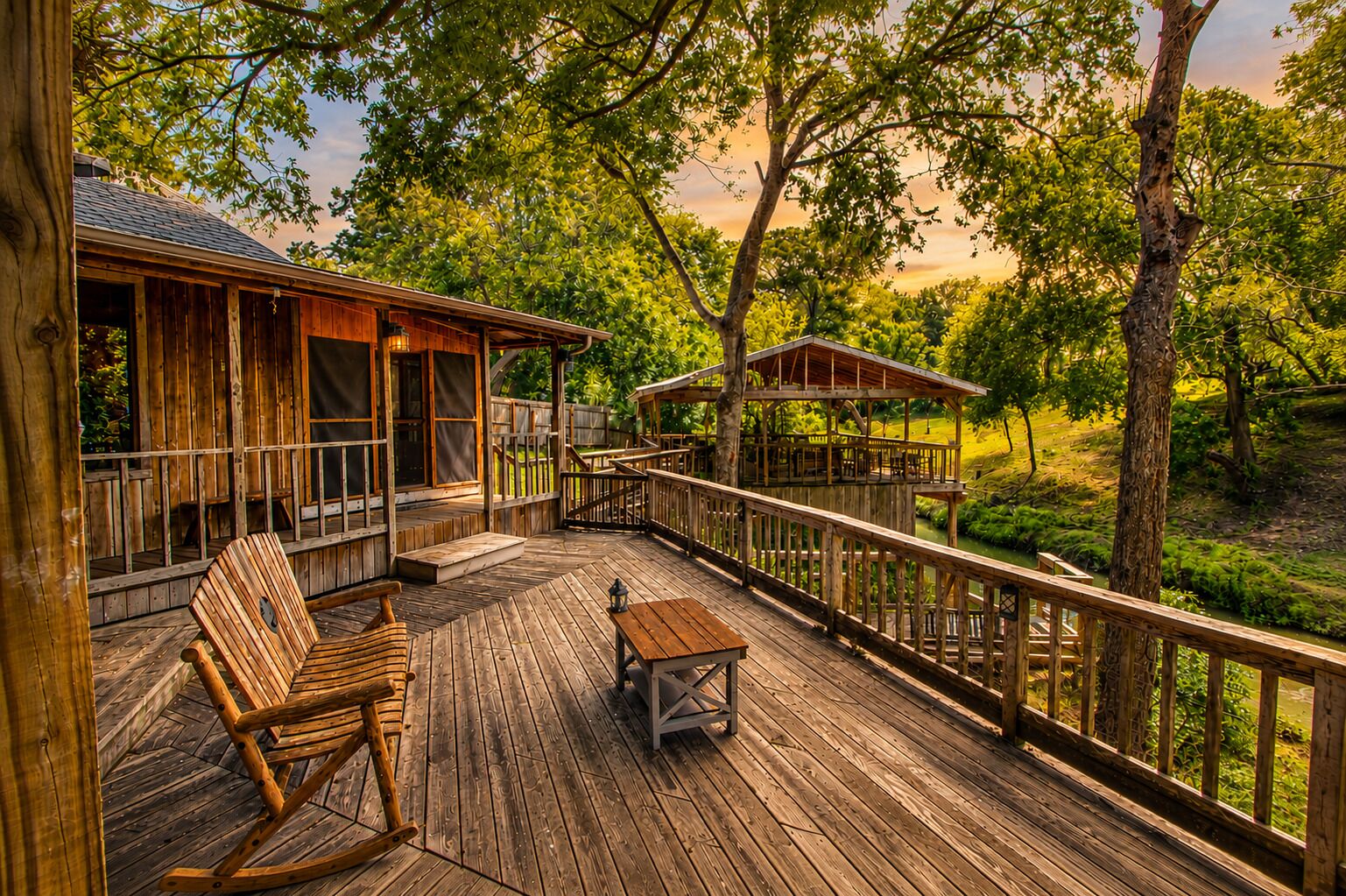 A wooden deck with a rocking chair and table, surrounded by trees and a view of a river.