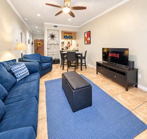A living room with a sofa, ottoman, TV, and a ceiling fan. - 2BR Condo W/ Pool, Hot Tub, Grills, Walk to Schlitterbahn! (New Braunfels)