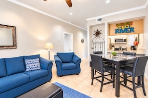 A living room with a blue sofa, a matching armchair, and a dining area with a table and chairs. - 2BR Condo W/ Pool, Hot Tub, Grills, Walk to Schlitterbahn! (New Braunfels)