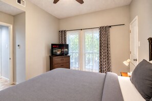 Une chambre à coucher avec un lit, un téléviseur posé sur une commode, une fenêtre avec des rideaux et une porte.