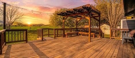 A wooden deck with a pergola, benches, and a view of the sunset over a field.