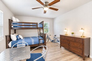 Un lit superposé avec un bureau et une chaise, une commode, un vélo et un ventilateur de plafond dans une pièce au plancher en bois et dotée d’une fenêtre.