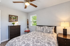 Ein Schlafzimmer mit Bett, Kommode, Stuhl, Nachttisch, Lampe, Deckenventilator, Fenster und einem gerahmten Bild an der Wand.