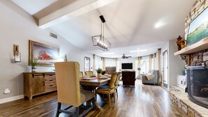 Un comedor amplio con una mesa de madera, sillas y una araña. Hay una chimenea con detalles de piedra y un televisor montado en la pared.