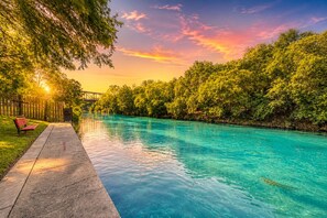 Una piscina con muelle de madera y un banco, rodeada de abundante vegetación y un cielo al atardecer.
