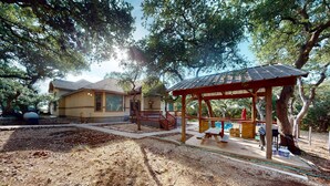 Un espacio al aire libre cubierto con mesas de picnic y una pérgola, rodeado de árboles y un edificio con techo inclinado.