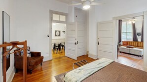 Un dormitorio con una cama, una silla de madera, una mecedora, un ventilador de techo y vistas a un salón con un sofá y una mesa.